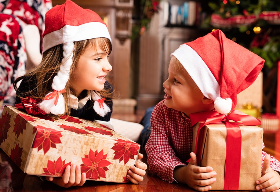 Две усмихнати деца с коледни шапки държат подаръци пред елхата по време на празничните приготовления.